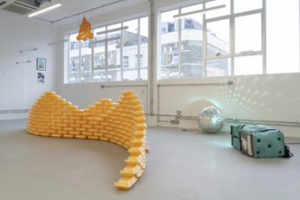 view of artwork in industrial-looking gallery interior. The artwork is a sculptural floorpiece, with a larger bright yellow element comprising geometric forms arranged in a large curving shape and to the right a silver sphere and a soft green and white cubic form