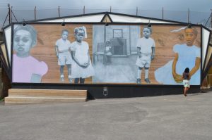photo view of a mural-like artwork, a large image running set before a wall, composed of collaged elements of mainly black and white images of children against a pale brown, woodgrain backdrop