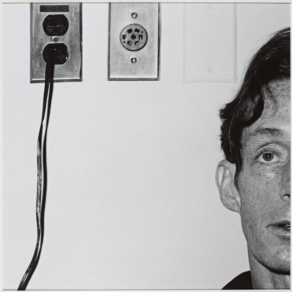 Photograph of John McKendry 1975, taken by Robert Mapplethorpe; the image is black and white, a man is staring ahead, and he is next to electrical plug sockets.