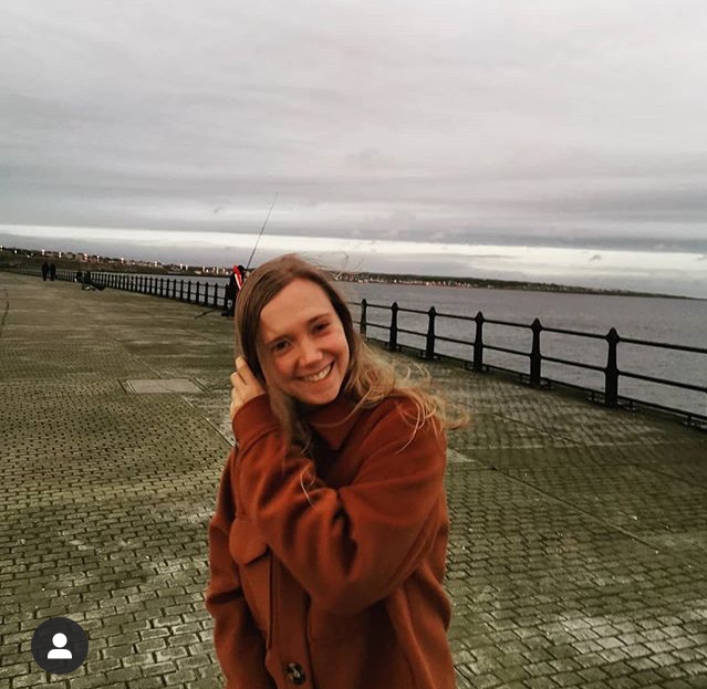 A photograph of Jess Starns who is standing on a promenade by the sea. She is looking directly into camera.