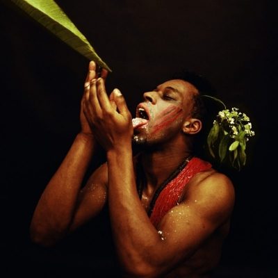 a man reaches up to drink off a leaf, against a stark black background