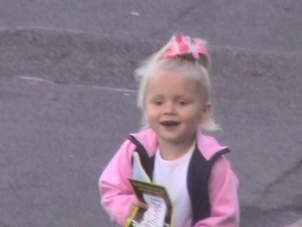 still from a video of a child in a pink jacket smiling, a concrete surface behind