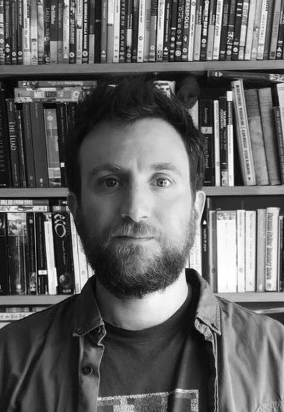 black and white portrait photo of Ben looking directly at camera, head and shoulders, casually dressed, bookshelves filled behind