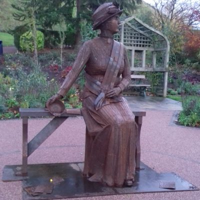 photograph of a brown metal statue in a park, a figure of a Edwardian woman in a dress seate