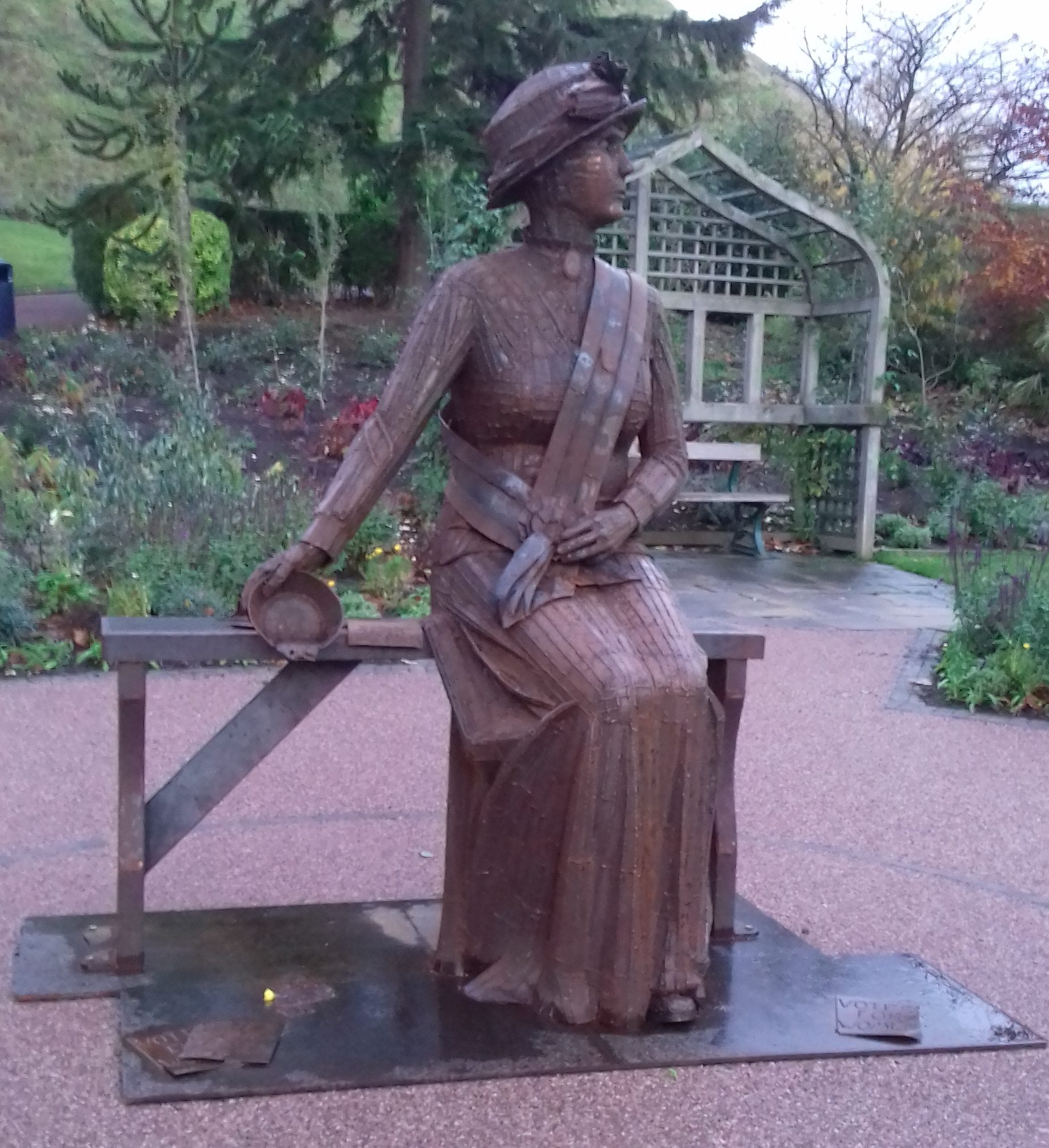 photograph of a brown metal statue in a park, a figure of a Edwardian woman in a dress seate