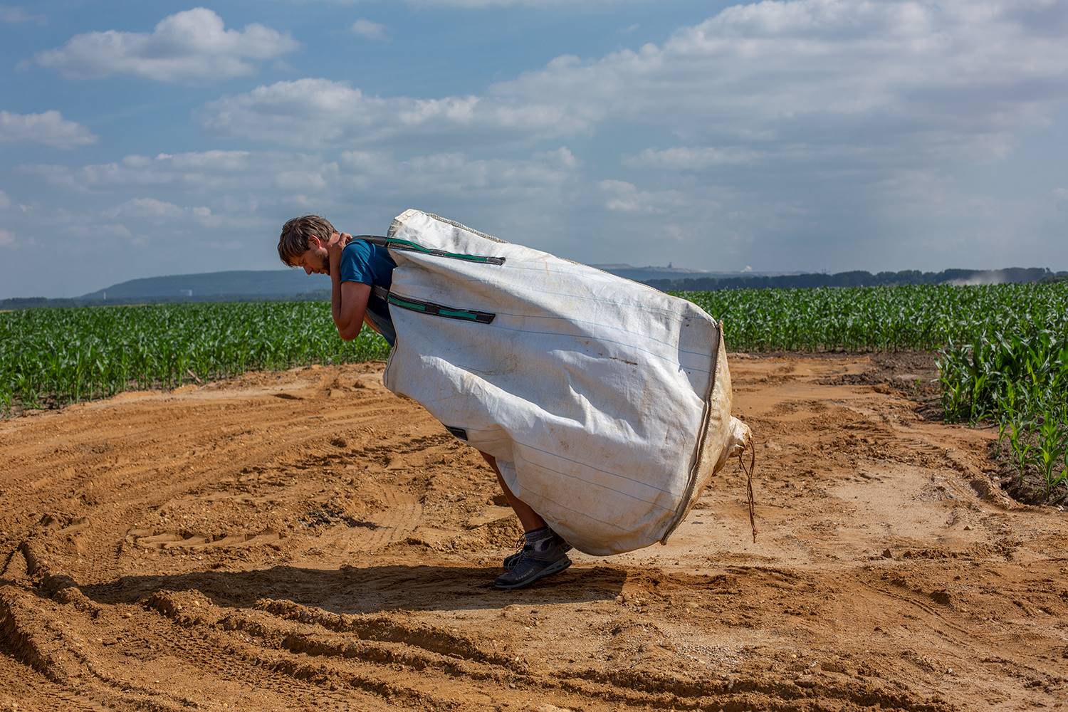 photographic view of a man hauling a huge white sack on his back, going from right to left across an open field
