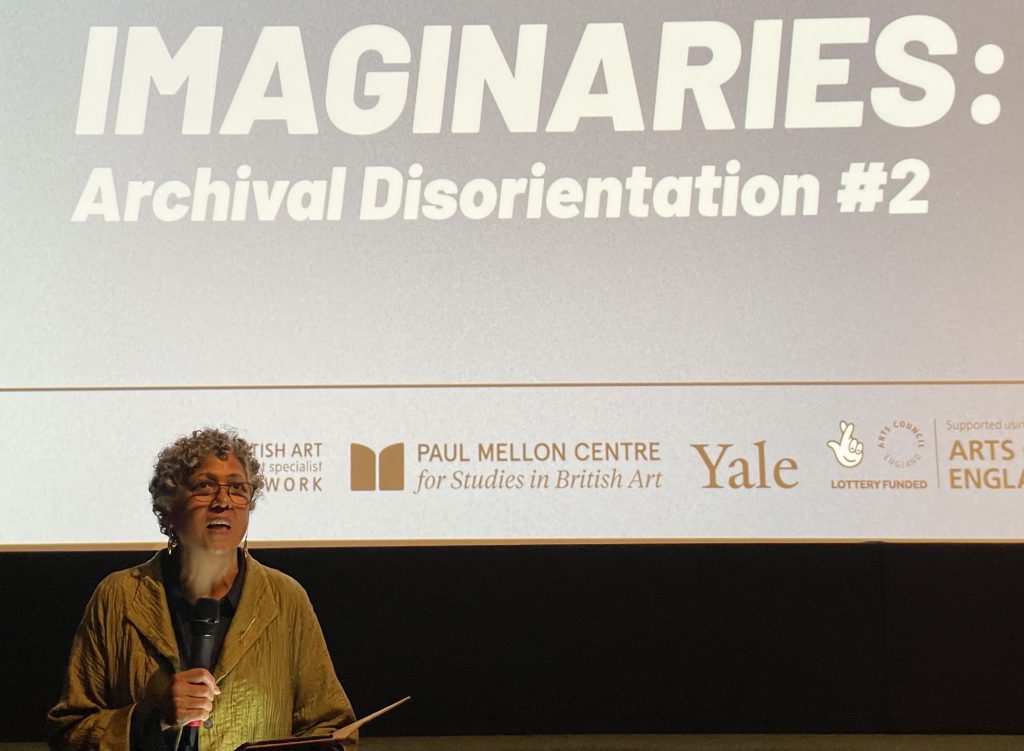 photo of Roshini Kempadoo talking, in front of a cinema screen with a slide announcing 'Itinerant Imaginaries: Archival Disorientation"
