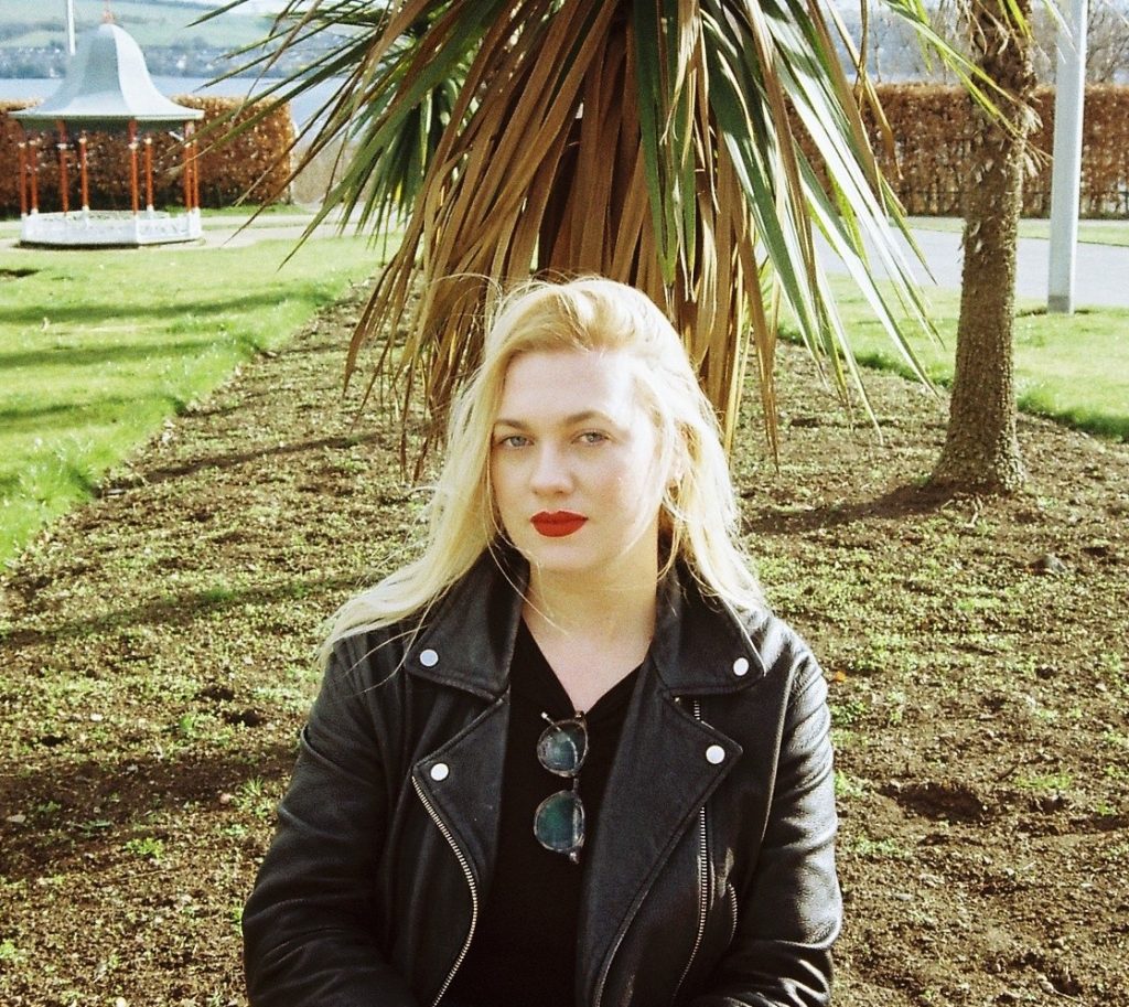 Laura is standing in front of a palm tree wearing a leather jacket.