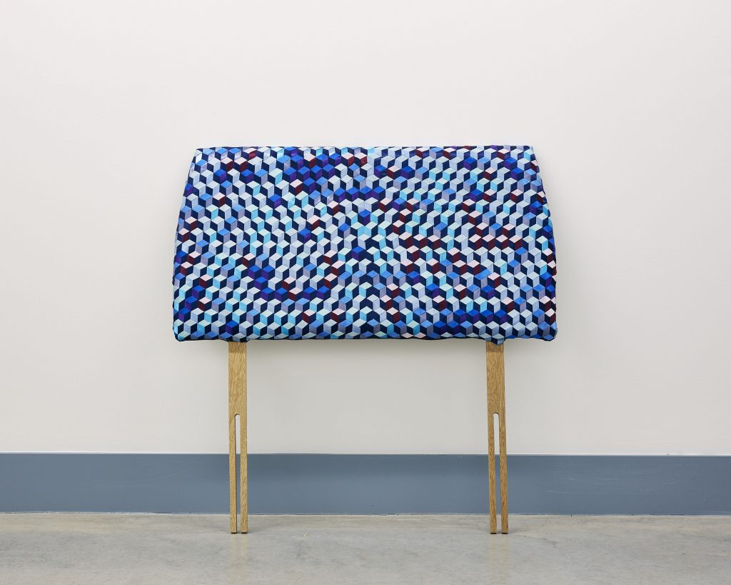 view of artwork, a headboard leaning against a grey gallery wall, the headboard covered in blue fabric featuring a geometric pattern