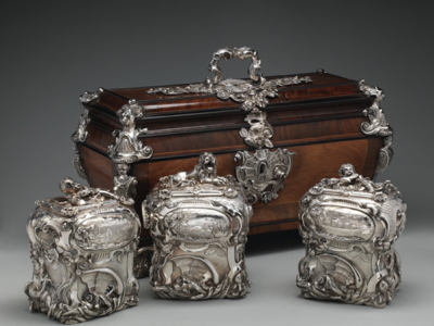 photograph in a neutral setting of a set of ornate silver objects, including a wooden case with silver fittings and three silver cases, all richly decorated in rococo style