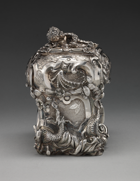 photograph in a neutral setting of a silver sugar box, ornately decorated with curvy organic designs