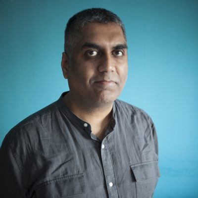 portrait photograph of Ashok Mistry wearing a grey collarless shirt in front of a plain blue background