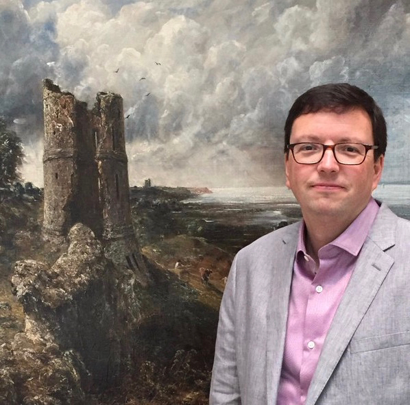 portrait photograph of Tim Barringer in grey jacket and pink shirt, standing in front of John Constable's painting of Hadleigh Castle, a stormy ruin scene