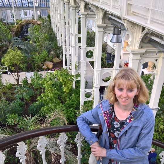 portrait photograph of Caitlin Doley standing on an ironwork balcony, green trees and a white ironwork industrial structure behind them