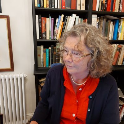 portrait photograph of Caroline Dakers sitting, before a bookcase loaded with books