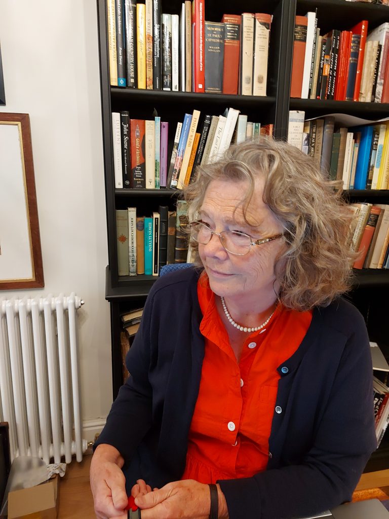 portrait photograph of Caroline Dakers sitting, before a bookcase loaded with books