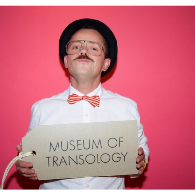 photo portrait of EJ Scott in white suit with red and white stripeybowtie and black hat, posed as if in a criminal headshot against a red background, holding a sign reading 'Museum of Transology'