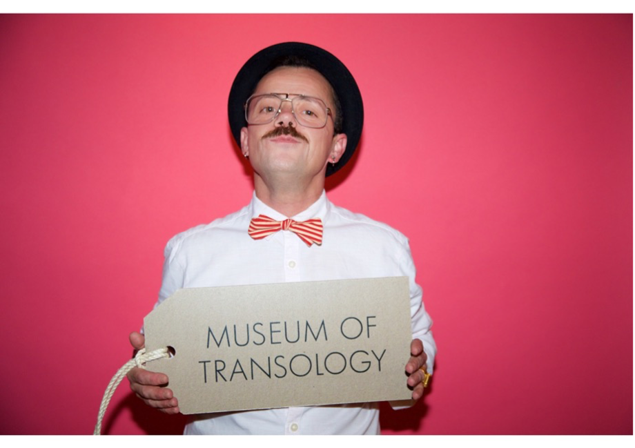 photo portrait of EJ Scott in white suit with red and white stripeybowtie and black hat, posed as if in a criminal headshot against a red background, holding a sign reading 'Museum of Transology'