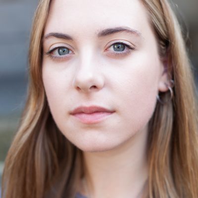 portrait photograph of Holly looking directly at the camera, in close up, a blurred architecttural background