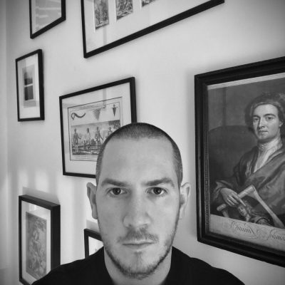 black and white photographic image of Peter Moore, in an interior with framed works of art behind