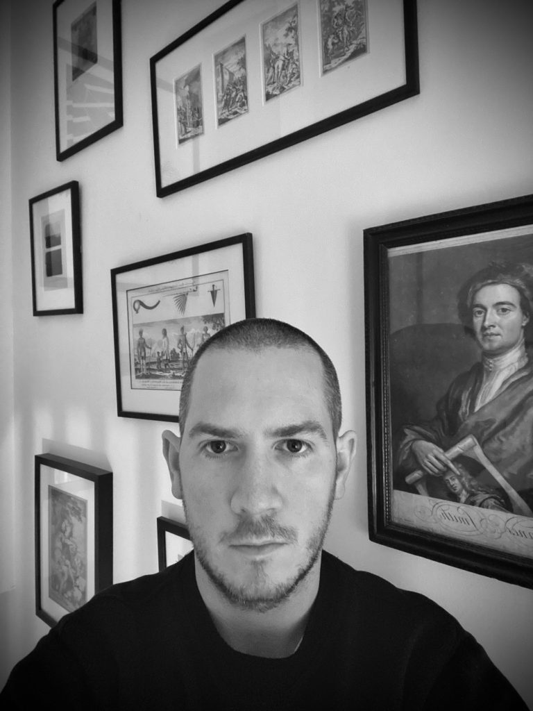 black and white photographic image of Peter Moore, in an interior with framed works of art behind
