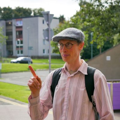 portrait photo of Matthew standing outside, with a backpack and flat cap, pointing; in the background a modern building suggestive of a university campus