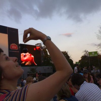 image of a crowd at an evening outdoor event; in the background a large screen with an image of a female figure dropping a cherry into their mouth; in the foreground the same image is recreated in person