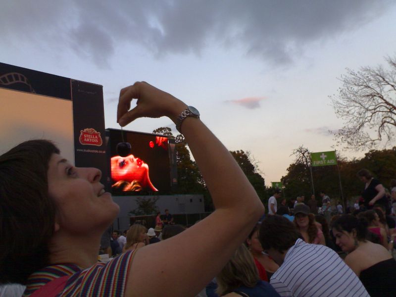 image of a crowd at an evening outdoor event; in the background a large screen with an image of a female figure dropping a cherry into their mouth; in the foreground the same image is recreated in person