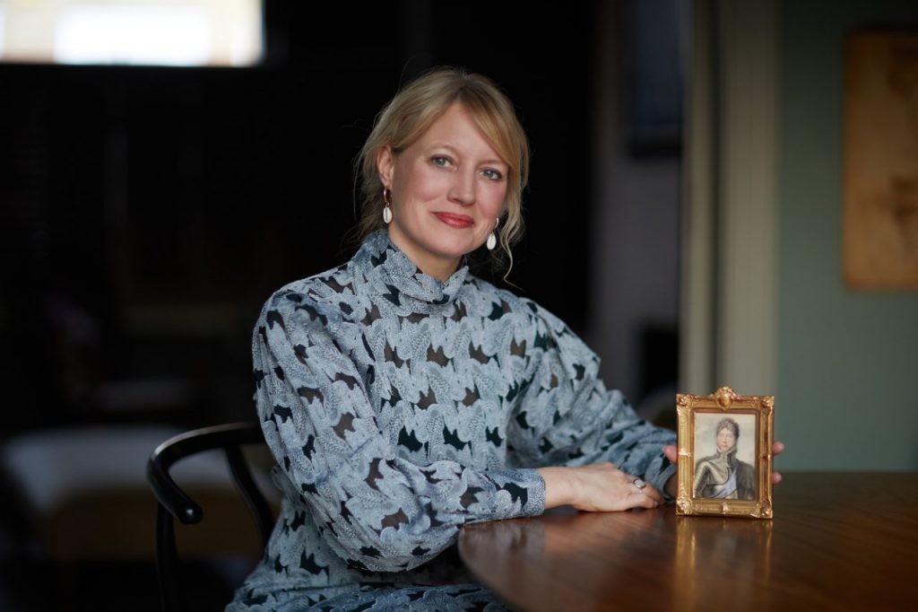 portrait photo of Emma seated at a table, a portrait miniature upright on the table in front of her