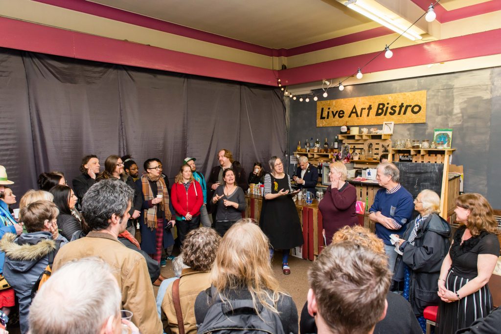 a crowd in a large room, a grey curtain covering the walls, a sign, 'Live Art Bistro' at back, in the centre Gill doing a speech, the crowd in a circle around