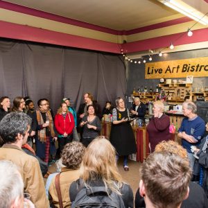 a crowd in a large room, a grey curtain covering the walls, a sign, 'Live Art Bistro' at back, in the centre Gill doing a speech, the crowd in a circle around
