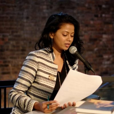 photo of Grace Aneiza Ali, leaning forward towards a microphone on a stand, a book on the table in front of them, papers in hand, in the act of speaking