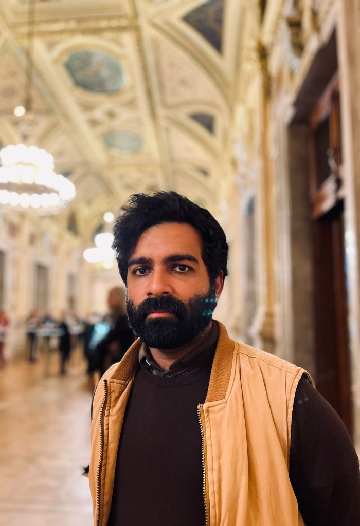 half length photo portrait of Hassan looking into camera, an architectural interior behind, a long corridor space with ornate ceiling decoration