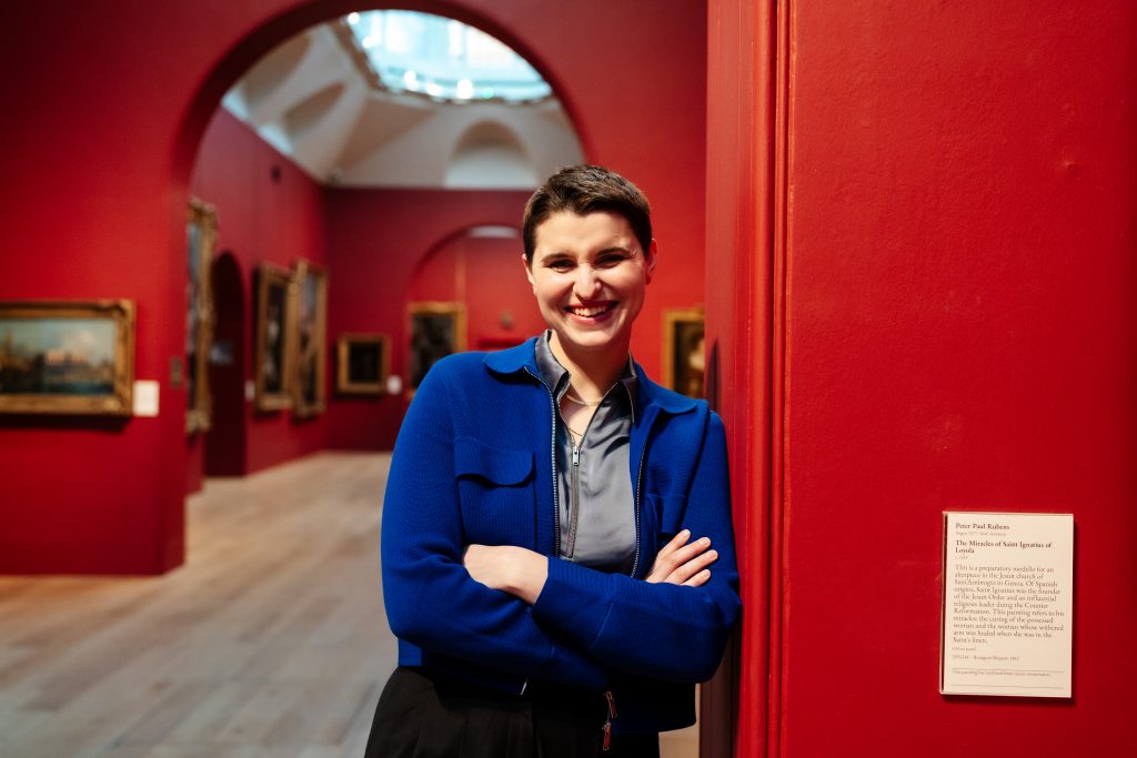 photo portrait of Helen standing, leaning against a wall in the interior of Dulwich Picture Gallery, the walls of the gallery a rich red, a skylight visible behind