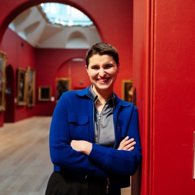photo portrait of Helen standing, leaning against a wall in the interior of Dulwich Picture Gallery, the walls of the gallery a rich red, a skylight visible behind