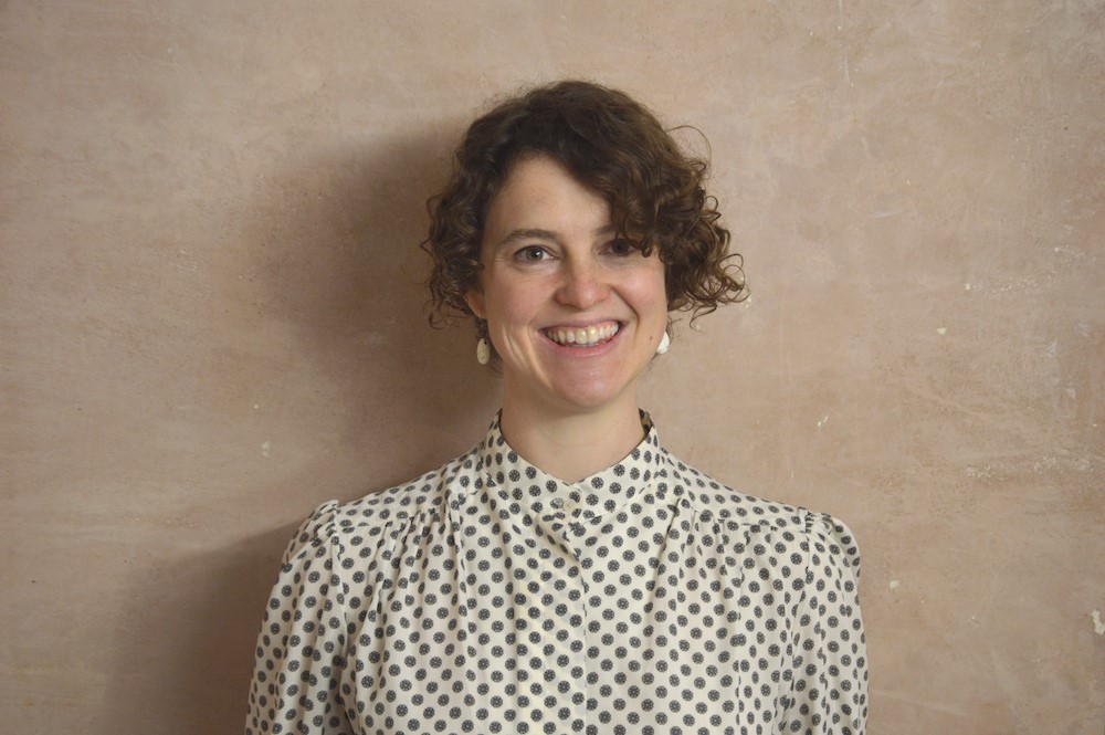 portrait photo of Kate, smiling, stood against pale brown wall