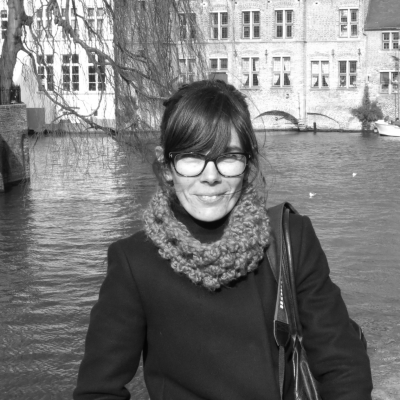 black and white image of Luisa, in outdoor coat, half length in front of water with waterside buildings in the distance behind