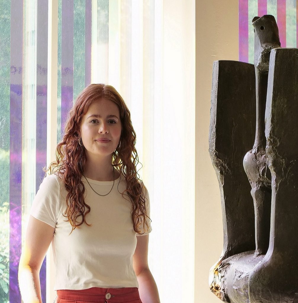 portrait photo of Sophie, half-length, standing in a brightly daylit gallery with a dark abstract sculptural form to the right