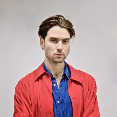 portrait photo of Will in a strongly coloured red jacket and blue shirt, head and shoulders with a plain grey background