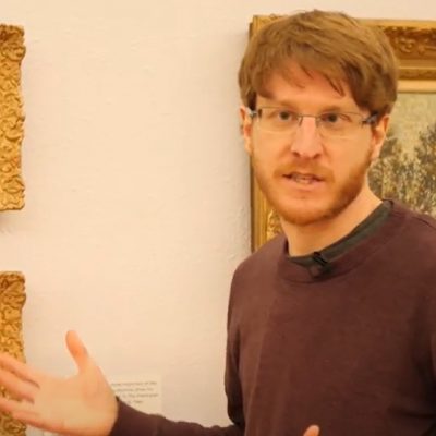 photo of Samuel in the act of speaking, right hand raised, in front of a wall with gilt framed paintings