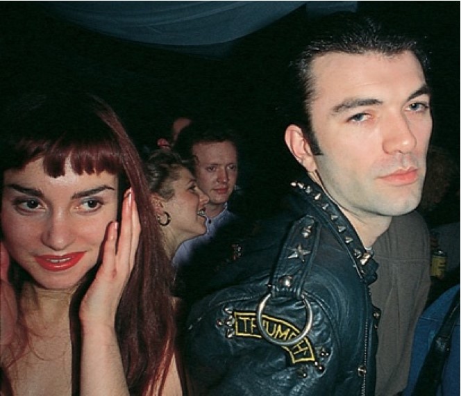 photograph, a crowded club - a female figure to the left in makeup, hand to face, a male figure with leather jacket to the right, looking out of shot