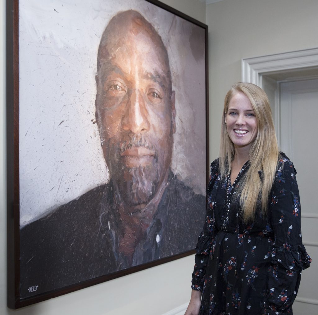 photo of Charlotte standing in a gallery space, smiling, a monumental portrait image of the cricketer Viv Richards to the left