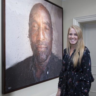 photo of Charlotte standing in a gallery space, smiling, a monumental portrait image of the cricketer Viv Richards to the left