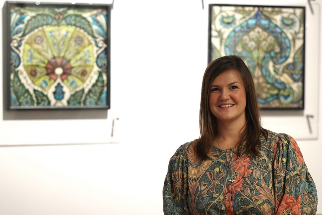 half length image of Sarah standing, smiling, in a gallery interior, decorative squares (De Morgan designs?) behind