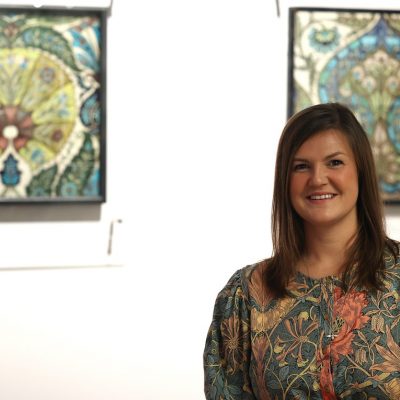 half length image of Sarah standing, smiling, in a gallery interior, decorative squares (De Morgan designs?) behind