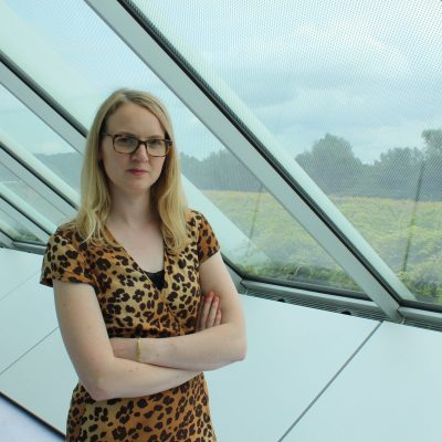 half length portrait photo of Tania, standing cross armed in a glass and metal interior (The Sainsbury Centre) view of grass and distant trees beyond