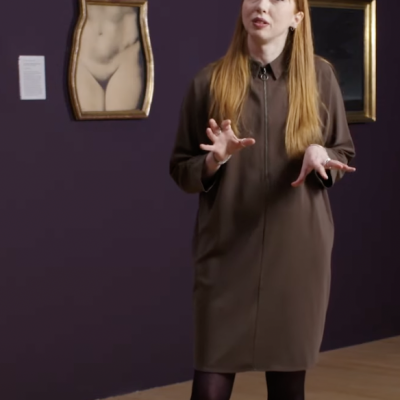 photo of Tor standing, stalking in a darkened art gallery setting, gesturing with both hands, a picture of a female figure's waist and pudenda in a shaped frame to the left