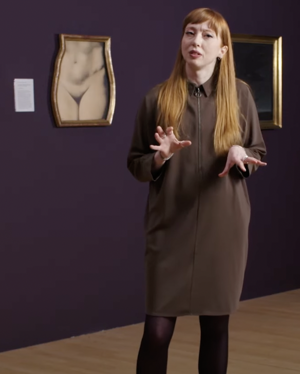 photo of Tor standing, stalking in a darkened art gallery setting, gesturing with both hands, a picture of a female figure's waist and pudenda in a shaped frame to the left
