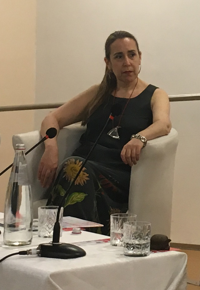 photo of Valeria seated in chair, turned to one wide, a table with papers and microphone in front, suggesting a seminar setting