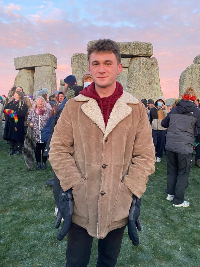 three quarter length portrait photo of Adan, standing with crowd of figures behind, Stonehenge in background